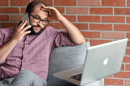 An employee using a phone while looking frustrated at his laptop