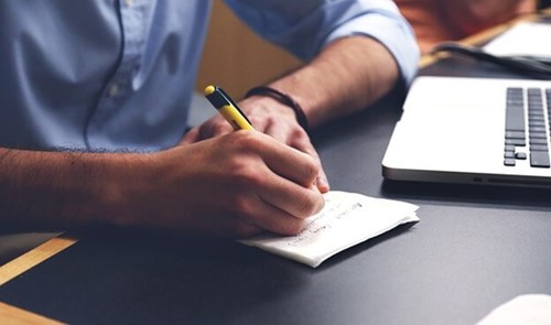 man writing on a notepad in front of a laptop